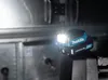 A Makita LED battery-powered flashlight is attached to a metal pipe in a dimly lit industrial or workshop setting. Produktbild-Vorschau