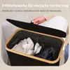 A person's hand is shown lifting the lid of a black laundry basket with a wooden frame, revealing laundry inside. Produktbild-Vorschau