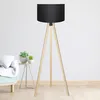 A floor lamp with a black shade and wooden tripod legs is shown in a room with a sofa and a small table with plants. Anteprima dell’immagine prodotto