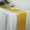 A gold satin table runner is laid across a white tablecloth, with a small vase of flowers and a geometric candle holder as decoration. Anteprima dell’immagine prodotto
