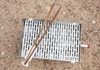 A metal grill plate with wooden tongs on a gravel surface. Produktbild-Vorschau