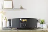 A black DAB radio with a color display is placed on a wooden desk next to a notebook, pencils, and a vase with dried flowers. A power outlet and a plant are visible in the background. Produktbild-Vorschau