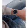 A person wearing a watch on their wrist, with denim jeans and a light-colored hoodie visible. Produktbild-Vorschau