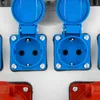 A close-up view of several electrical outlets, some blue and some red, mounted on a white surface. Produktbild-Vorschau