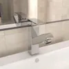 A close-up view of a modern chrome faucet installed in a white sink, with tiled walls in the background. Podgląd zdjęcia produktu