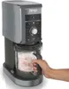 A hand is shown holding the container of an ice cream maker. The ice cream maker is on a white and gray surface. Podgląd zdjęcia produktu