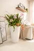 A white planter with a large arrangement of orchids is placed on a light wooden floor next to a framed artwork. In the background, a sofa with a beige throw blanket is visible, and a wooden shelf with decorative items is mounted on the wall. Náhled obrázku produktu