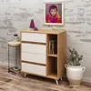 A dresser with white drawers and a wooden frame is placed against a textured white brick wall. A framed picture of a child is hung above the dresser. A small side table with a plant is to the left, and a large potted cactus is to the right. Produktbild-Vorschau