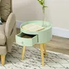 A light green round side table with an open drawer sits next to a beige sofa. A book and a vase with a flower are on top of the table. The table has wooden legs and is on a patterned rug. Voir les images du produit