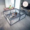 A baby is playing in a playpen in a living room, with a woman reading on a sofa in the background. Produktbild-Vorschau