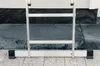 The bottom section of an aluminum ladder is shown resting on a tiled floor against a dark stone wall. Podgląd zdjęcia produktu