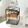 A modern side table with a glass top and a wood drawer is placed next to a white sofa, with a lamp and a vase of plants on top. Produktbild-Vorschau