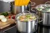 Three stainless steel pots are on a stovetop, one filled with spaghetti, another with vegetables, and the third with broccoli. Ingredients like onions, tomatoes, and parsley are on a wooden cutting board nearby. Produktbild-Vorschau