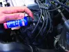 A hand is holding a small blue bottle and applying its contents to a part of a car engine. Produktbild-Vorschau