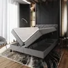 A modern bedroom with a grey upholstered bed frame, the mattress is lifted to reveal storage compartments underneath. The room features dark walls, a large window with white curtains, and decorative shelves with vases. Produktbild-Vorschau