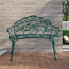 A green metal garden bench with a rose pattern is shown on a wooden deck in front of a textured wall. The background also shows a white lattice fence, green grass, and trees. Podgląd zdjęcia produktu