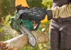 A person wearing protective gloves and clothing is using a green chainsaw to cut a tree branch outdoors. Produktbild-Vorschau