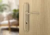 A close-up of a bronze door handle on a wooden door, with a blurred interior scene in the background. Produktbild-Vorschau