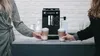 Two women are holding cups of coffee in front of a coffee machine. Produktbild-Vorschau