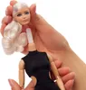 A doll's head with blonde hair is being held by hands, detached from the doll's body which is wearing a black dress. Náhled obrázku produktu