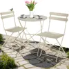 A small round table and two chairs set up on a stone patio with flowers and a bird.