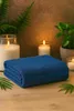 A folded blue towel is placed on a wooden surface, surrounded by lit candles and greenery, creating a spa-like atmosphere. Produktbild-Vorschau