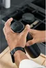 A person's hands are shown operating a black pepper mill over a wooden cutting board, with a stovetop visible in the background. Podgląd zdjęcia produktu