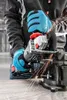 A person wearing blue gloves is using a cordless angle grinder to cut a metal rod clamped in a vise. Sparks are flying from the cutting action. Podgląd zdjęcia produktu