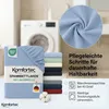 A folded bedsheet is shown in the foreground, with a stack of other colored bedsheets behind it. A washing machine is visible in the background. Produktbild-Vorschau