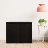 A black fabric laundry basket with a lid is placed in a room with a white wall and wooden floor. A small side table with a plant is to the right, and a pink pendant light hangs from the ceiling. Produktbild-Vorschau