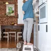 A person is standing on a white folding stool in a kitchen, reaching into a cabinet. Produktbild-Vorschau