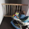 A baby in a walker is near a wooden baby gate at the top of stairs. Voir les images du produit
