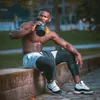 A muscular man sitting on a stone ledge and drinking from a water bottle. Produktbild-Vorschau