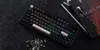 A black mechanical keyboard with RGB lighting is placed on a dark textured surface next to two books and a bar spoon. Produktbild-Vorschau
