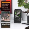 A Brandson CO2 measurement device is shown on a desk next to a plant, with a woman working on a laptop in the background. Produktbild-Vorschau