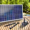 A solar panel, a water pump, and two attachments are displayed on a wooden deck next to a pond. Produktbild-Vorschau