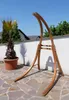 A wooden hammock stand is displayed outdoors on a stone patio, with a potted palm tree and a white wall in the background. Produktbild-Vorschau