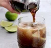 A hand is pouring a dark liquid from a glass bottle into a glass filled with ice and lime slices. The drink is fizzy and appears to be a cola. Produktbild-Vorschau