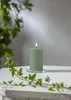 A green LED pillar candle with a lit flame sits on a white surface, with ivy vines in the foreground and background. Podgląd zdjęcia produktu