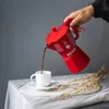A hand pours coffee from a red Moka pot into a white cup on a table covered with a white cloth. Anteprima dell’immagine prodotto