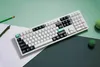 A white mechanical keyboard is placed on a surface with a light green and dark green background, along with two pencils and two green balls. Voir les images du produit