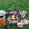 An outdoor cooking setup with a tripod stand holding a grill over a campfire, surrounded by firewood, a pot of soup, and ingredients for cooking. Produktbild-Vorschau