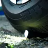 A flashlight is placed on the ground next to a car tire, with the flashlight beam illuminating the tire and the ground. Anteprima dell’immagine prodotto