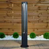 A modern black bollard light stands on a paved patio in front of a wooden fence, with decorative white stones and green spherical bushes. Anteprima dell’immagine prodotto