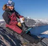 A woman is sitting on a rock in a mountainous, snowy landscape, putting on a ski boot. She is wearing winter clothing and goggles. Voir les images du produit