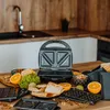 A sandwich maker and various accessories, including different plates and ingredients like oranges and broccoli, are displayed on a wooden table in a kitchen setting. Anteprima dell’immagine prodotto