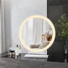 A round makeup mirror with LED lighting is placed on a dark countertop, with some cosmetic products and a vase with reeds to the left, and a potted plant to the right. Podgląd zdjęcia produktu