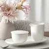 A white ceramic creamer and sugar bowl with lid are placed on a white oval tray, next to a vase with pink flowers. Náhled obrázku produktu