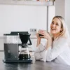 A woman is smiling and holding a cup of coffee next to a coffee maker. Produktbild-Vorschau