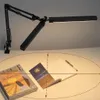 A black LED architect lamp is shown clamped to a desk, illuminating a stack of magazines and an open book. Náhľad obrázku produktu